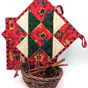 Puede incluir: Dos agarraderas de Navidad acolchadas con ribete a cuadros rojos y negros. Las agarraderas presentan un diseño de patchwork con hombres de jengibre, estrellas y lazos sobre un fondo rojo. Una pequeña cesta de mimbre contiene palitos de canela y bayas rojas.