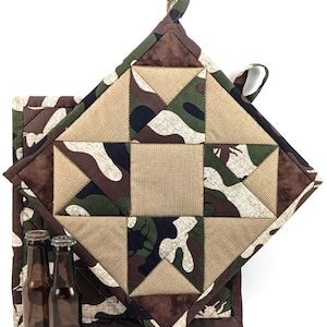 Puede incluir: Dos agarraderas de olla acolchadas en un patrón de camuflaje con tela marrón, verde y beige. Las agarraderas tienen un asa de bucle.