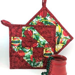 May include: Two red quilted pot holders with a Christmas ornament and pine bough pattern. The pot holders have a loop for hanging. A strand of green beads and a red ceramic mug are in the foreground.