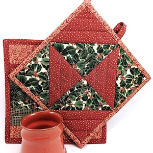Puede incluir: Dos agarraderas de olla acolchadas rojas con un patrón de acebo y bayas. Las agarraderas tienen un ribete dorado y un lazo para colgar. Una taza de cerámica roja y cascabeles dorados están en primer plano.