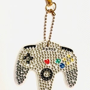 May include: A gold chain keychain with a white, rhinestone-covered video game controller charm. The controller has black buttons and a colorful button pad.