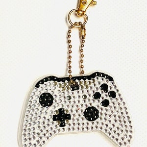 May include: A white and black video game controller keychain with rhinestones. The keychain has a gold chain and a gold clasp.