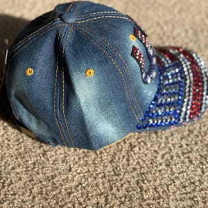 May include: A denim baseball cap with a red, white, and blue American flag design on the back. The flag is embellished with rhinestones.