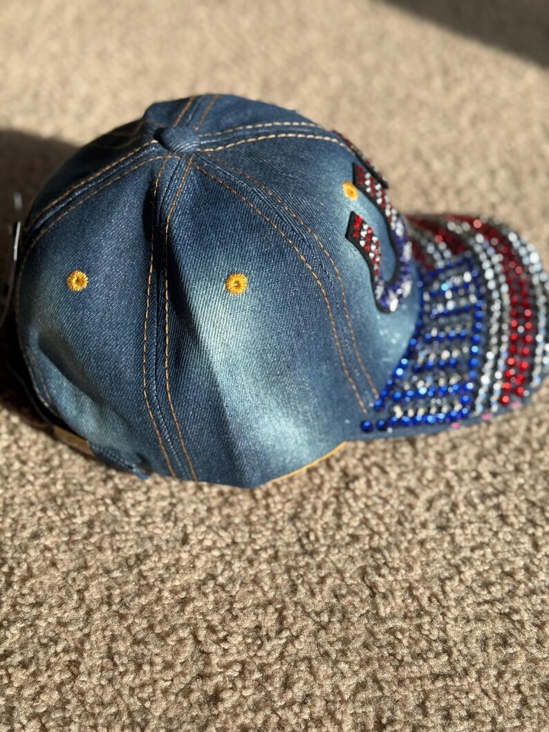 May include: A denim baseball cap with a red, white, and blue American flag design on the back. The flag is embellished with rhinestones.
