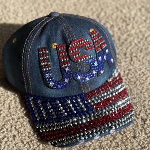 May include: A denim baseball cap with a red, white, and blue American flag design. The flag is made of rhinestones and the word "USA" is spelled out in rhinestones on the front of the cap.