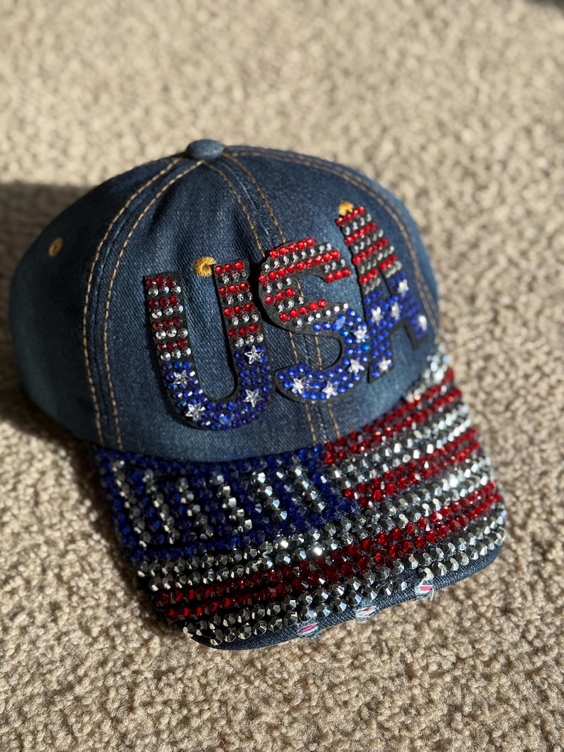 May include: A denim baseball cap with a red, white, and blue American flag design. The flag is made of rhinestones and the word "USA" is spelled out in rhinestones on the front of the cap.