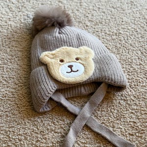 Op de afbeelding: Een taupe gebreide baby muts met een lichtbruine teddybeer gezicht applicatie. De muts heeft een bijpassende pompon bovenop en lange banden. De muts is gemaakt van een zacht, gebreid materiaal.