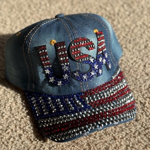 May include: A denim baseball cap with a red, white, and blue American flag design. The flag is made of rhinestones and the letters "USA" are also made of rhinestones.