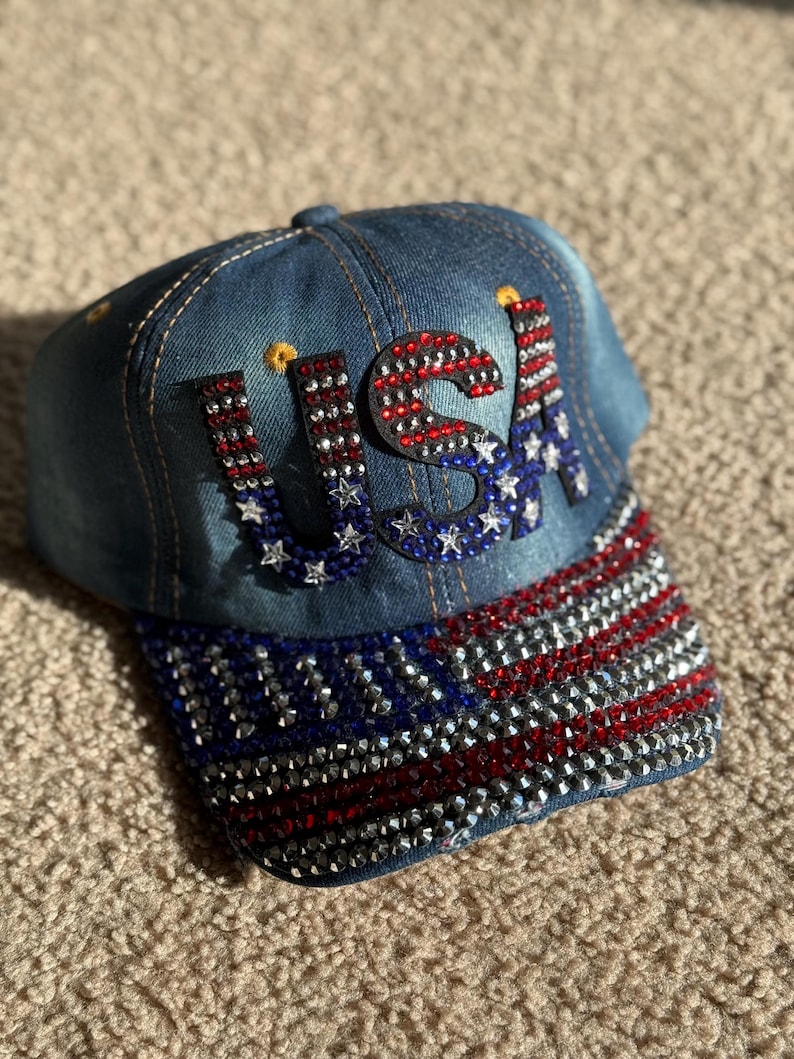 May include: A denim baseball cap with a red, white, and blue American flag design. The flag is made of rhinestones and the letters "USA" are also made of rhinestones.