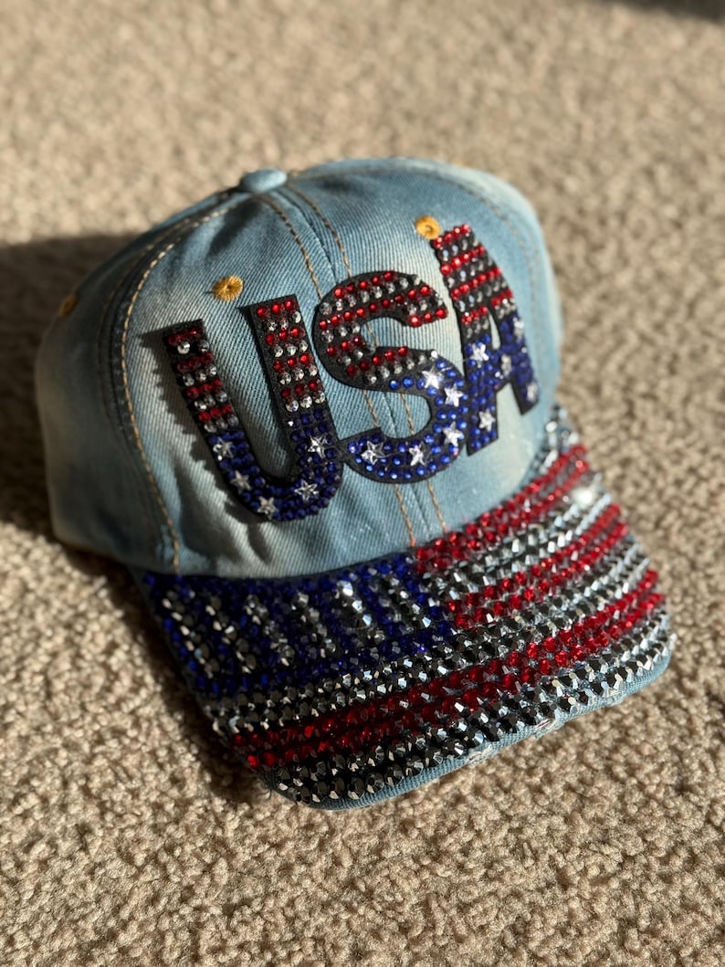 May include: A blue denim baseball cap with a red, white, and blue rhinestone 'USA' design on the front and a matching American flag design on the brim.