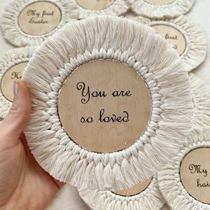 May include: Set of round wooden coasters with white fringe and a variety of engraved messages, including "My first Easter", "My first Thanksgiving", "My first Christmas", "You are so loved", and "My first birthday".