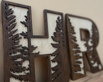 May include: Wooden letters spelling "HR" with a wood grain pattern and a cutout design of trees. The letters are brown with a light wood background.