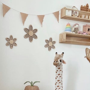 May include: A white wall with a brown wooden shelf with a rainbow stacking toy, a stuffed bunny, and a small pink toy car. There are also three wooden flower decorations on the wall, a stuffed giraffe, and a butterfly sticker. The shelf has the word "BABY" on it.