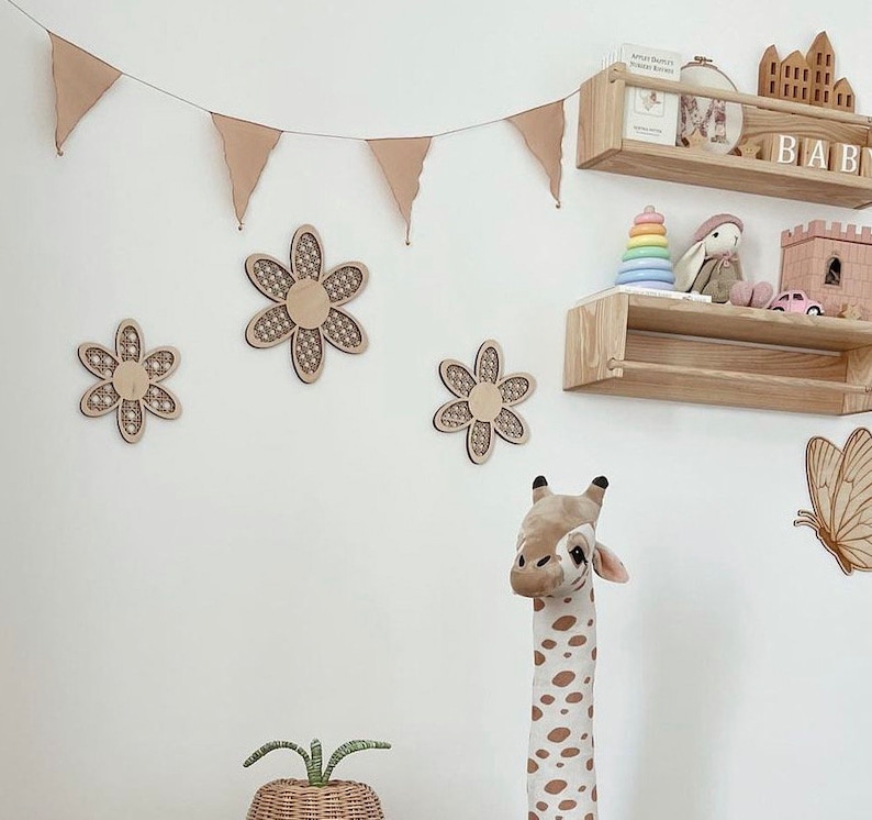 May include: A white wall with a brown wooden shelf with a rainbow stacking toy, a stuffed bunny, and a small pink toy car. There are also three wooden flower decorations on the wall, a stuffed giraffe, and a butterfly sticker. The shelf has the word "BABY" on it.