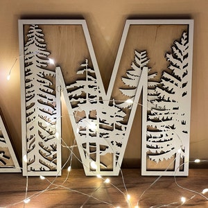 May include: Wooden letter "M" with a white frame and a natural wood backing. The letter features a forest scene cut-out design. A string of small white lights is draped in front of the letter, creating a warm glow. The letter is displayed on a wooden surface.