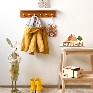 Puede incluir: Un perchero de madera con ganchos, una chaqueta amarilla colgada, un conejito de madera con el nombre "Emily" en él, una cordillera de madera con el nombre "Ethan" en él, una figurita de conejito de madera y dos botas de lluvia amarillas.