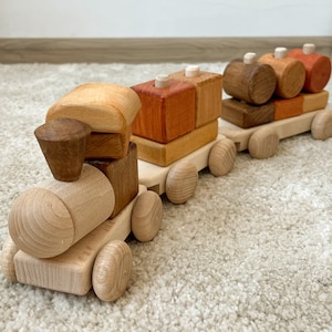 May include: A wooden toy train with colorful blocks as cars. The train is made of natural wood and has a brown, orange, and red color scheme. The train is on a light beige carpet.