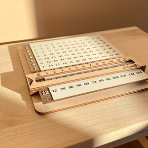 May include: A wooden multiplication chart with a sliding ruler. The chart features numbers 1 through 12 on the left side and numbers 12 through 144 on the bottom. The sliding ruler is labeled with numbers 12, 24, 36, 48, 60, 72, 84, 96, 108, 124, 132, and 144.