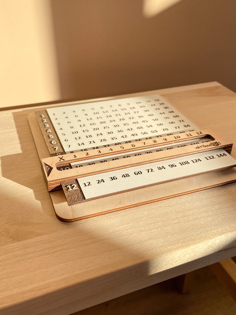 May include: A wooden multiplication chart with a sliding ruler. The chart features numbers 1 through 12 on the left side and numbers 12 through 144 on the bottom. The sliding ruler is labeled with numbers 12, 24, 36, 48, 60, 72, 84, 96, 108, 124, 132, and 144.