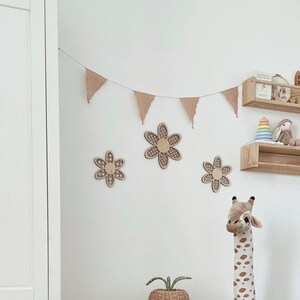 May include: A nursery room decorated in a neutral color scheme with wooden shelves, a wooden giraffe toy, and a wooden flower garland. The shelves are decorated with wooden toys, including a castle, a bunny, and a rainbow stacking toy. The garland is made of wooden flowers with a brown and white pattern. The room is decorated with a neutral color scheme, with white walls and a white closet.