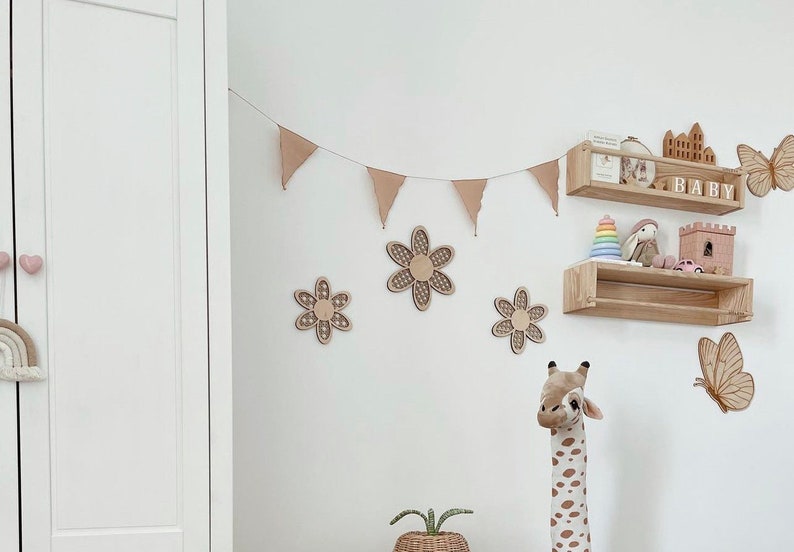 May include: A nursery room decorated in a neutral color scheme with wooden shelves, a wooden giraffe toy, and a wooden flower garland. The shelves are decorated with wooden toys, including a castle, a bunny, and a rainbow stacking toy. The garland is made of wooden flowers with a brown and white pattern. The room is decorated with a neutral color scheme, with white walls and a white closet.