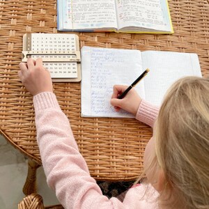 May include: A young person sits at a table with a wooden multiplication chart and a notebook. They are writing with a pen in the notebook. The notebook has a grid pattern and the multiplication chart has numbers 1 through 100.
