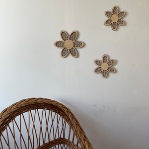 May include: Three wooden wall decor pieces in the shape of a flower with a woven pattern. The flowers are light brown and are arranged in a triangular pattern on a white wall. A wicker chair is in the foreground.