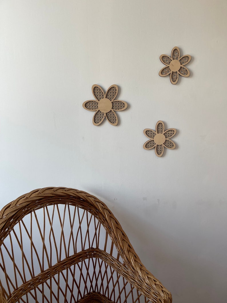 May include: Three wooden wall decor pieces in the shape of a flower with a woven pattern. The flowers are light brown and are arranged in a triangular pattern on a white wall. A wicker chair is in the foreground.