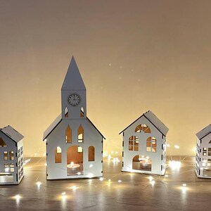 Può includere: Cinque case in legno bianco con finestre e porte ritagliate, illuminate da candele votive. Le case sono disposte in fila su una superficie in legno con luci di fata bianche.