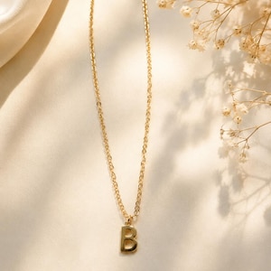 Puede incluir: Un collar de tono dorado que presenta una cadena delicada y un colgante único con la forma de la letra B mayúscula. El colgante tiene un acabado liso y pulido, y está suspendido del centro de la fina cadena de eslabones metálicos entrelazados.