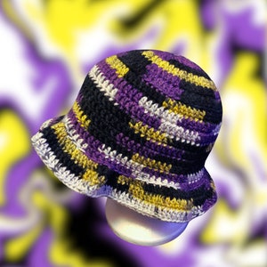 Könnte beinhalten: Ein handgehäkelter Fischerhut in den Farben der nicht-binären Pride-Flagge. Der Hut hat Streifen in Schwarz, Lila, Gelb und Weiß. Der Rand ist einfarbig weiß. Der Hut wird auf einer weißen, abgerundeten Form präsentiert.