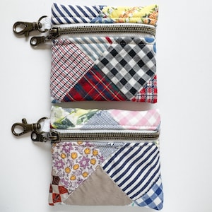 May include: Two small, patchwork fabric pouches with brass-colored zippers and clips. The pouches feature a variety of patterned fabrics, including stripes, plaid, and floral designs. The top pouch has a blue and white striped fabric at the top, and the bottom pouch has a blue and white striped fabric at the bottom.