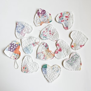 May include: A garland of twelve heart-shaped ornaments made from quilted fabric patches. Each heart features a unique combination of white fabric and various colorful floral or geometric patterns, all stitched together with a visible grid-like quilting texture.
