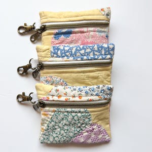 以下が含まれることがあります： パッチワーク生地で作られた小さなジッパー付きポーチが4つ。各ポーチは異なる花柄で、金属製のキーリングに取り付けられています。ポーチは互いに積み重ねられています。