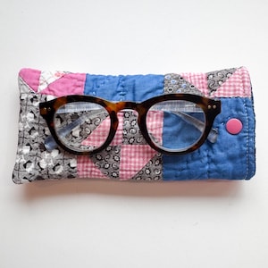 May include: A pair of tortoise shell glasses with round frames resting on a rectangular quilted fabric case. The case features a patchwork design of blue, pink, and black and white patterned fabric squares with a single pink snap button closure.