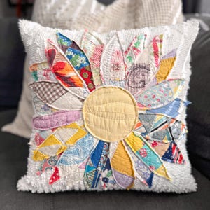 May include: A decorative square pillow with a large, colorful sunflower design. The sunflower petals are made from various fabric scraps in shades of red, blue, yellow, and green. The center is a solid yellow circle, and the pillow has a textured white background.
