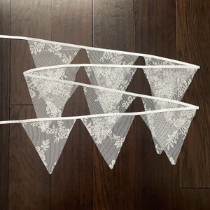 May include: A white lace bunting with triangular flags. The flags have a delicate floral pattern and are connected by white ribbon.