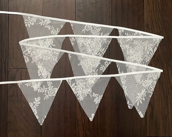 Könnte beinhalten: Eine weiße Spitzengirlande mit dreieckigen Flaggen. Die Flaggen haben ein zartes Blumenmuster und sind durch ein weißes Band verbunden.