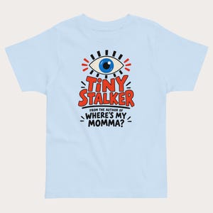 May include: A light blue t-shirt featuring a graphic of a blue eye with black lashes and red text that reads TINY STALKER. Below this, smaller black text reads FROM THE AUTHOR OF WHERE'S MY MOMMA? with decorative black lines surrounding the text.
