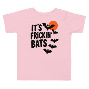May include: A light pink short-sleeve t-shirt featuring a graphic print with the black text "IT'S FRICKIN' BATS" alongside five black bat silhouettes and a solid orange circle representing a moon, all centered on the front of the garment.