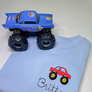 May include: A light blue t-shirt with a red monster truck embroidered on the front. The name "Britton" is embroidered below the truck.