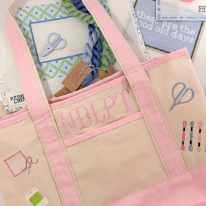 Op de afbeelding: Een beige draagtas met roze randen en handvatten, geborduurd met naaigerelateerde ontwerpen en de naam "NOLPT" in roze. De tas is gevuld met borduurgaren en borduurkaders. De achtergrond bevat borduurkaders met tekst.