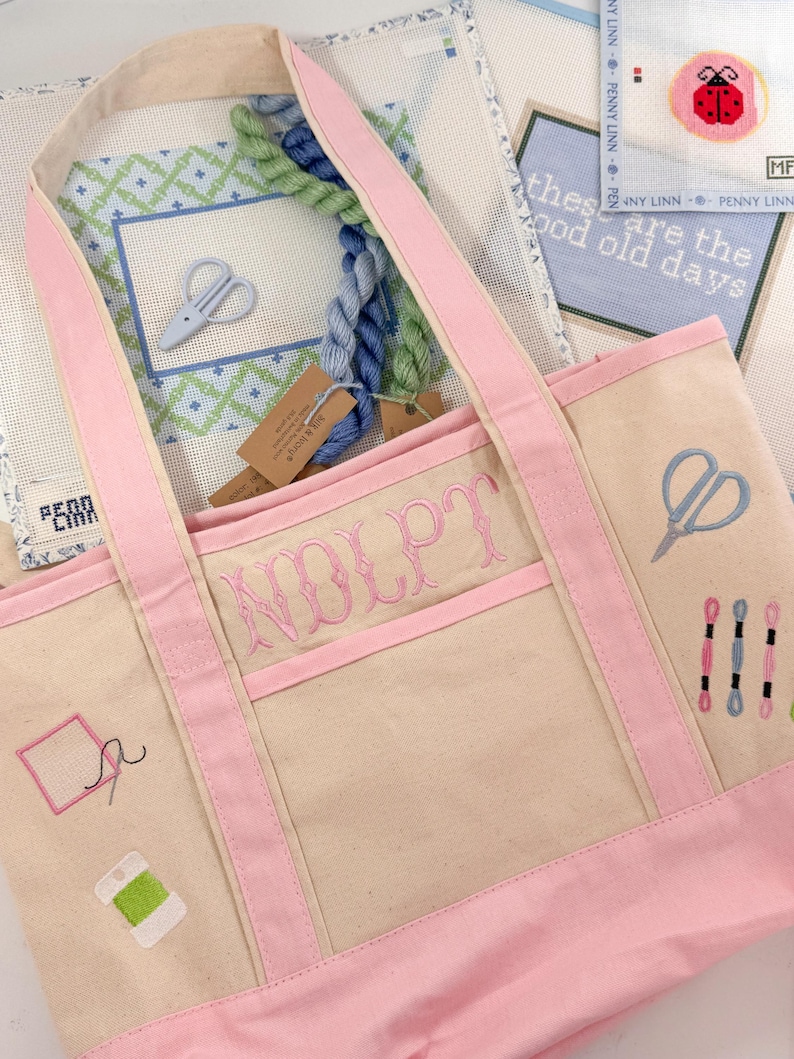 Op de afbeelding: Een beige draagtas met roze randen en handvatten, geborduurd met naaigerelateerde ontwerpen en de naam "NOLPT" in roze. De tas is gevuld met borduurgaren en borduurkaders. De achtergrond bevat borduurkaders met tekst.