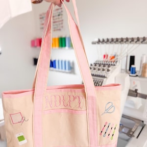 Op de afbeelding: Een beige en roze tote bag met geborduurde naaigerelateerde ontwerpen. De tas heeft een roze rand, handvatten en de initialen "NDLPT" geborduurd op de voorkant. Extra geborduurde elementen zijn scharen, klosjes garen en een naald en draad.