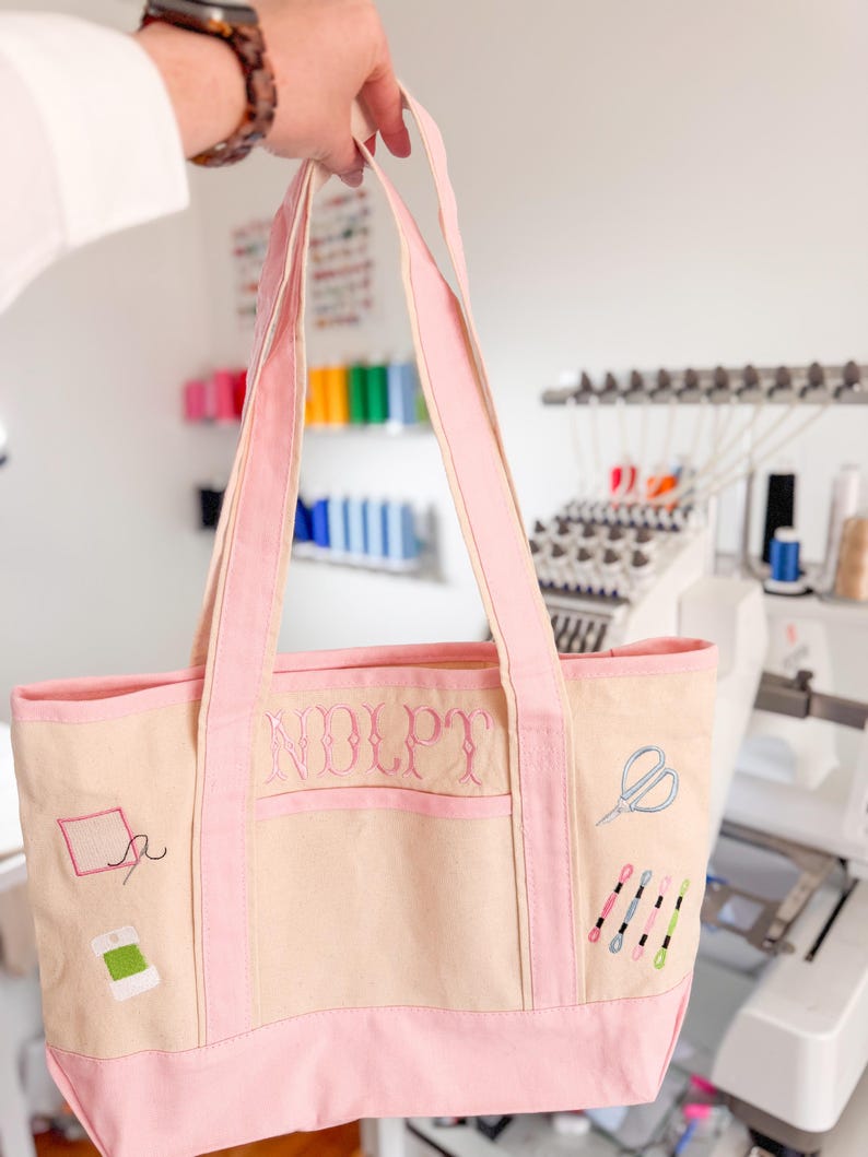 Op de afbeelding: Een beige en roze tote bag met geborduurde naaigerelateerde ontwerpen. De tas heeft een roze rand, handvatten en de initialen "NDLPT" geborduurd op de voorkant. Extra geborduurde elementen zijn scharen, klosjes garen en een naald en draad.