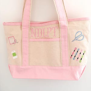 Op de afbeelding: Een beige canvas draagtas met roze accenten en borduursels met naaigerei. De tas heeft roze handvatten, een roze bodem en een roze zak. De voorkant van de tas heeft de geborduurde letters "NOLPT".