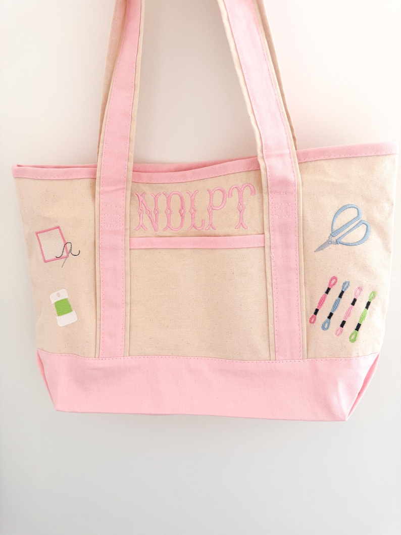 Op de afbeelding: Een beige canvas draagtas met roze accenten en borduursels met naaigerei. De tas heeft roze handvatten, een roze bodem en een roze zak. De voorkant van de tas heeft de geborduurde letters "NOLPT".