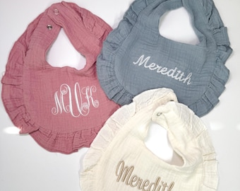 May include: Three ruffled baby bibs made of crinkled fabric. One is dusty rose with a white embroidered monogram, one is slate blue with white embroidered text reading Meredith, and one is cream with tan embroidered text reading Meredith.