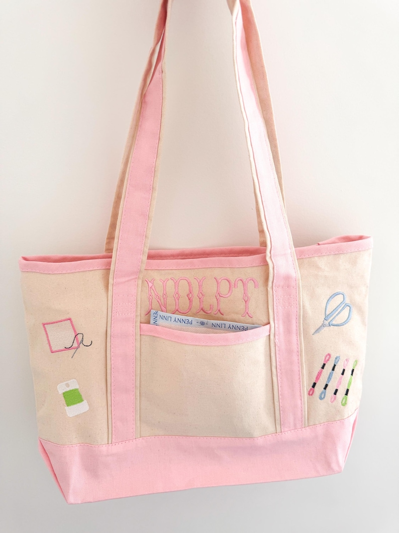 Op de afbeelding: Een lichtbeige draagtas met roze accenten, met geborduurde naaipatronen. De tas heeft een roze basis, rand en handvatten. Een voorvak toont de tekst "NDLPT" en een klein label. Naaigerei zoals scharen en garenspoelen zijn op de tas geborduurd.