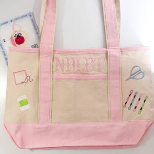 Op de afbeelding: Een beige canvas draagtas met roze accenten, met borduursels met naaigerei. De tas heeft roze handvatten en een roze bodem. De voorkant van de tas heeft de geborduurde letters "NOLPT". Extra geborduurde elementen zijn scharen, garenklosjes en een naald.